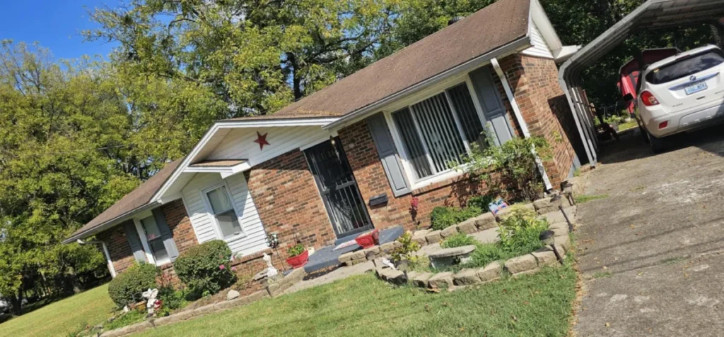 Single-story brick house with garden, front steps, and a driveway leading to a parked white SUV under a carport.