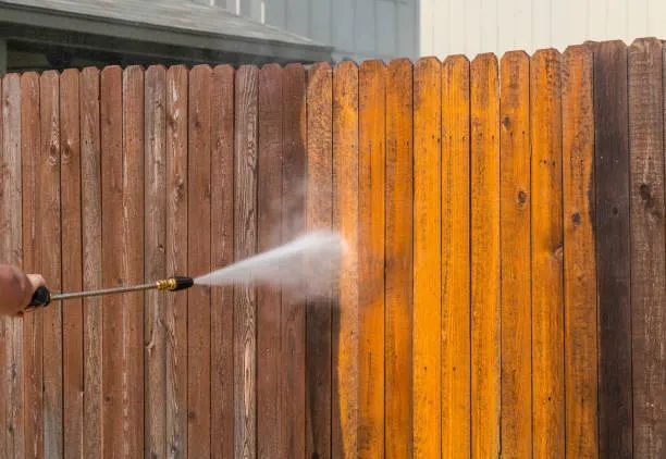A person power washes a wooden fence, revealing a cleaner, lighter surface compared to the untreated area.