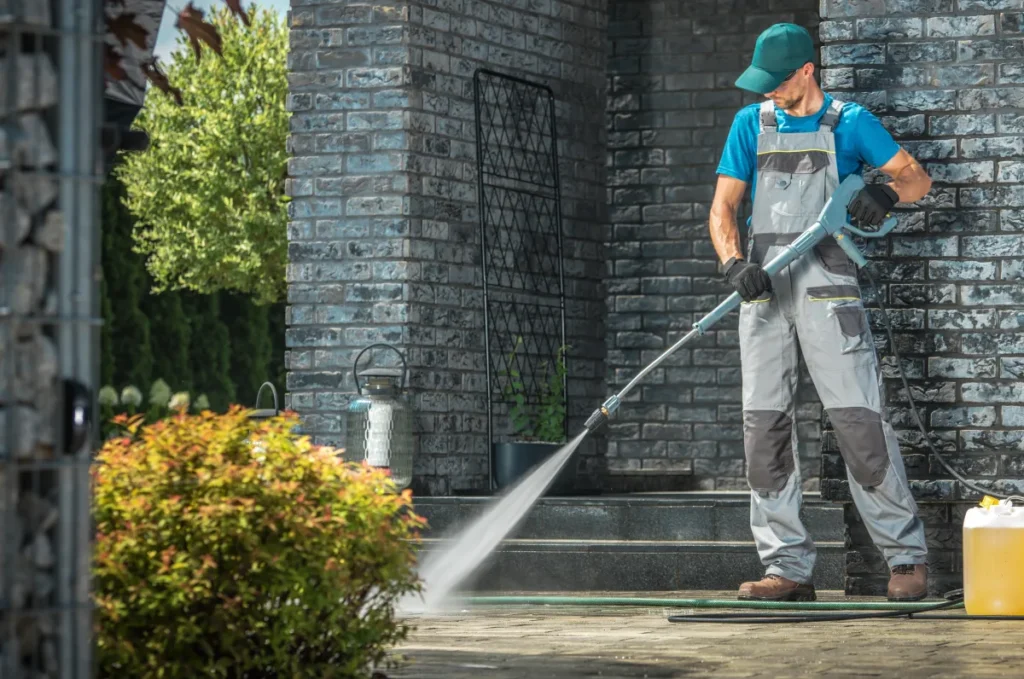 A worker in overalls uses a pressure washer to clean an outdoor stone walkway in front of a brick house.